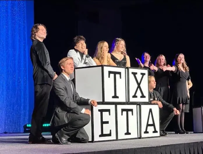 A lot of people standing on a stage with 5 boxes with the words txets